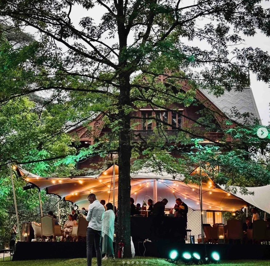 Outdoor evening event with a large white canopy tent, string lights, and guests seated and mingling under a large tree in front of a historic wooden house.