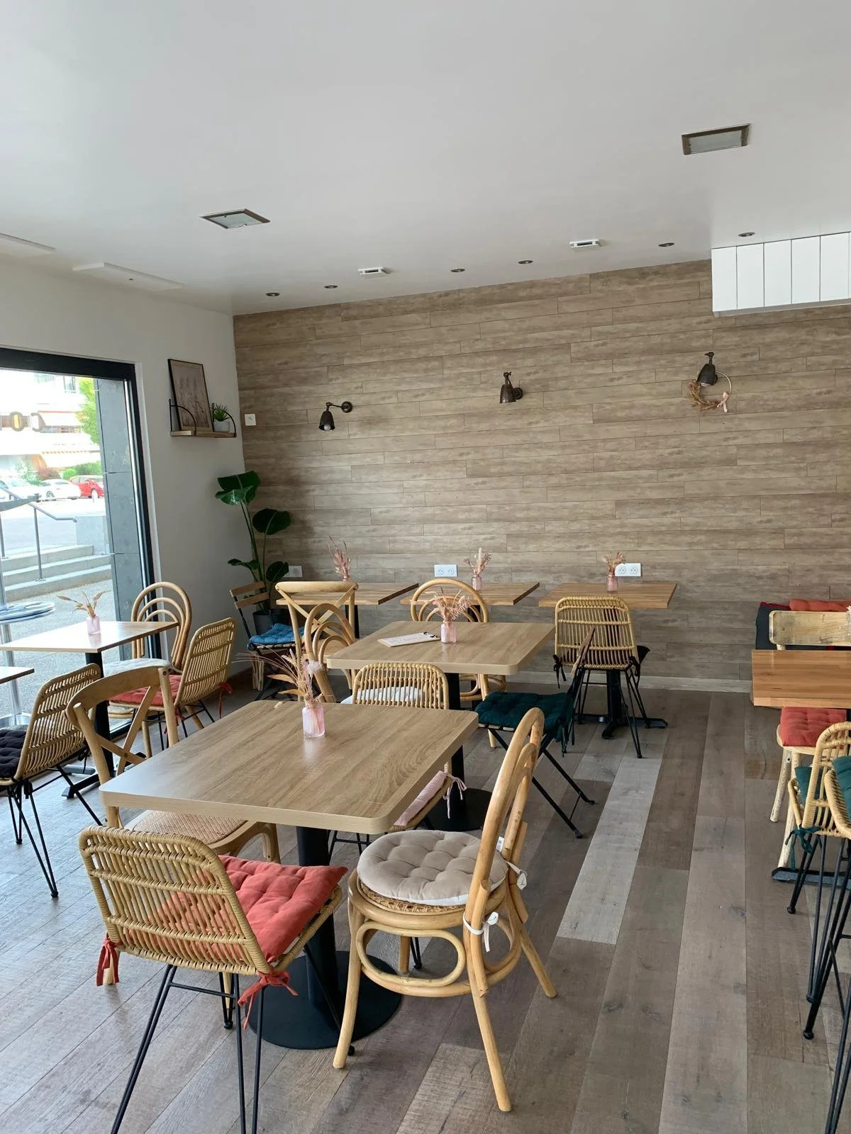 Empty cafe with wooden tables and chairs, decorated with pink vases and dried flowers, natural light from a glass door, beige wood wall, and wall-mounted reading lamps.