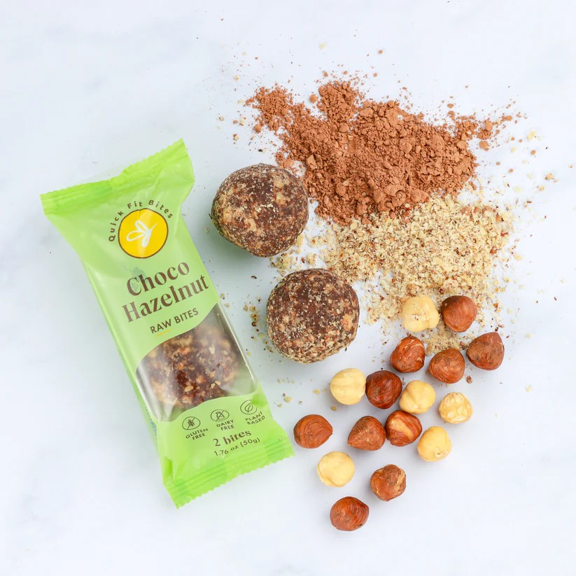 Choco hazelnut raw bites ingredients including hazelnuts, cocoa powder, crushed nuts, and two coated snack balls, some of which are in a green packet.
