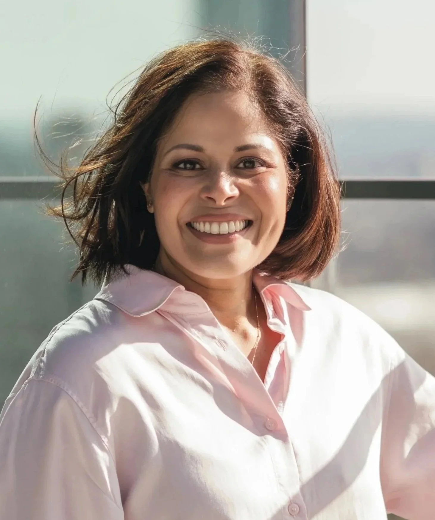 Smiling woman with shoulder-length brown hair, wearing a light pink button-up shirt, sitting near a large window with sunlight.