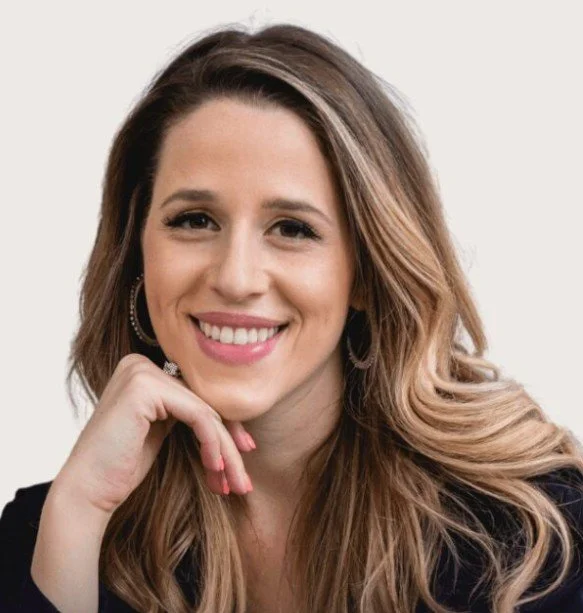 Young woman with long brown hair, wearing a white shirt and ripped jeans, sitting in a white room with decorative wall paneling, smiling at the camera.