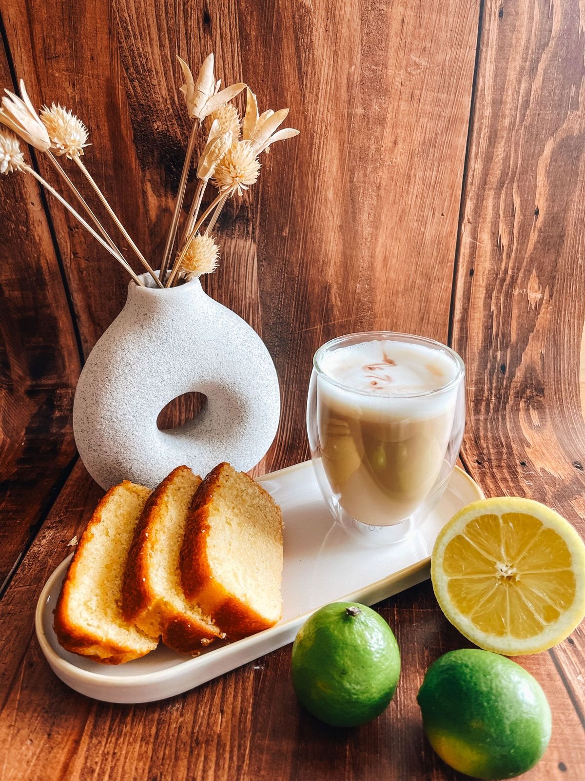 A still life of sliced cake, a glass of a creamy beverage, two lemons, two limes, a white ceramic vase with dried flowers, on a wooden surface and background.