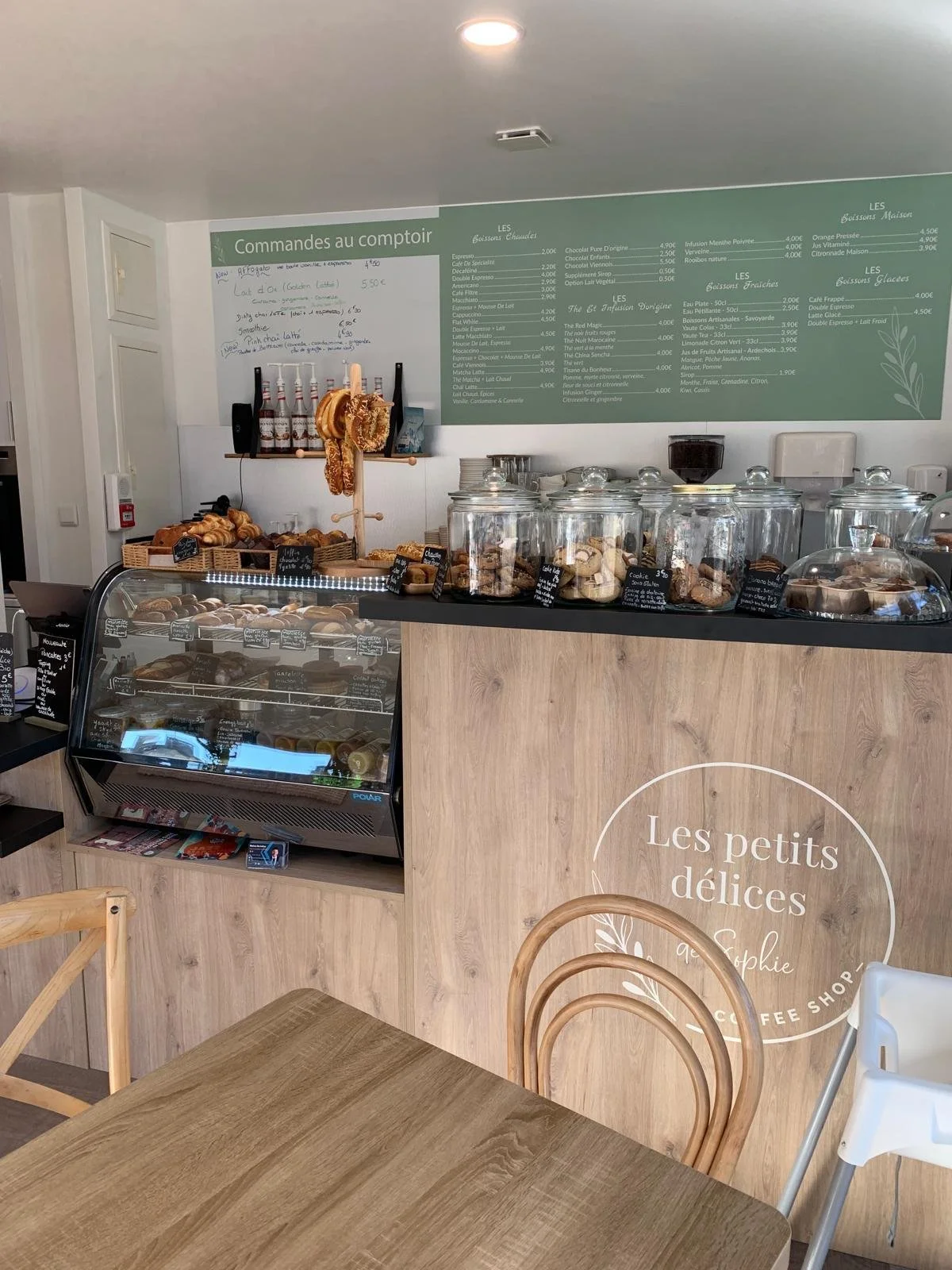 Inside a cozy coffee shop with a wooden counter displaying jars of baked goods such as cookies and pastries. There is a refrigerated display case filled with various desserts, a small table, and chairs. The background features a menu board with drink options and prices in French, and the shop's name, 'Les petits délices,' is written on the front of the counter.