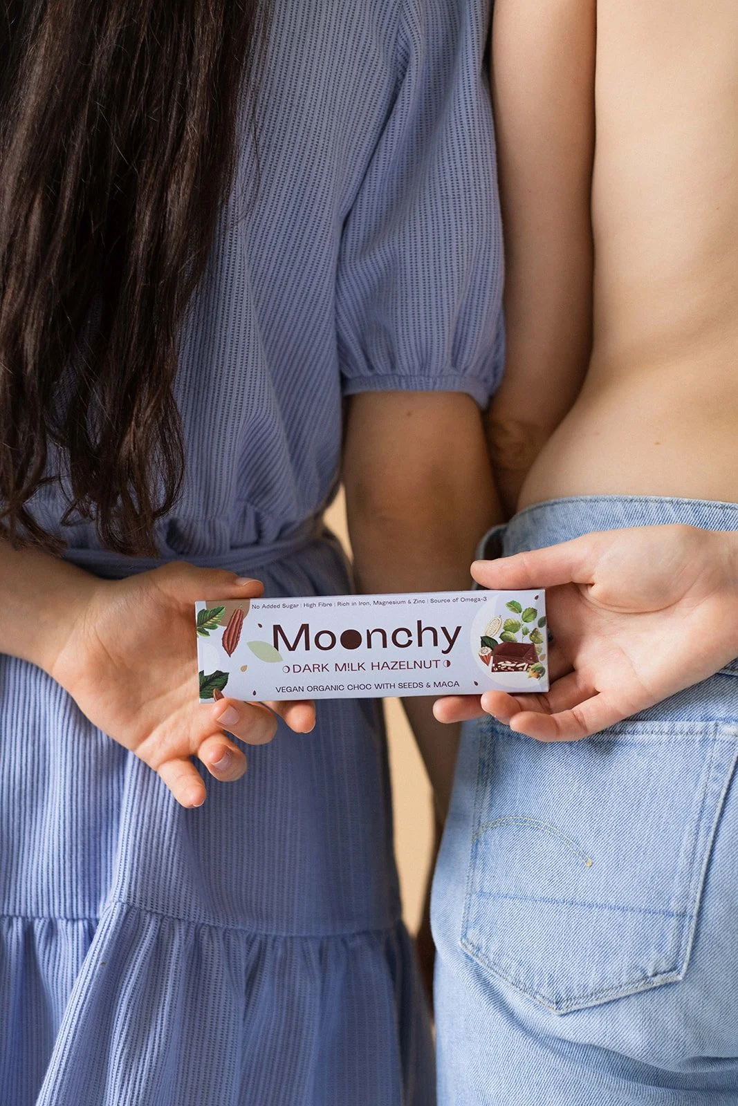 Two women hold a chocolate bar labeled 'Moonchy Dark Milk Hazelnut' between them, with one woman wearing a blue dress and the other shirtless with jeans.