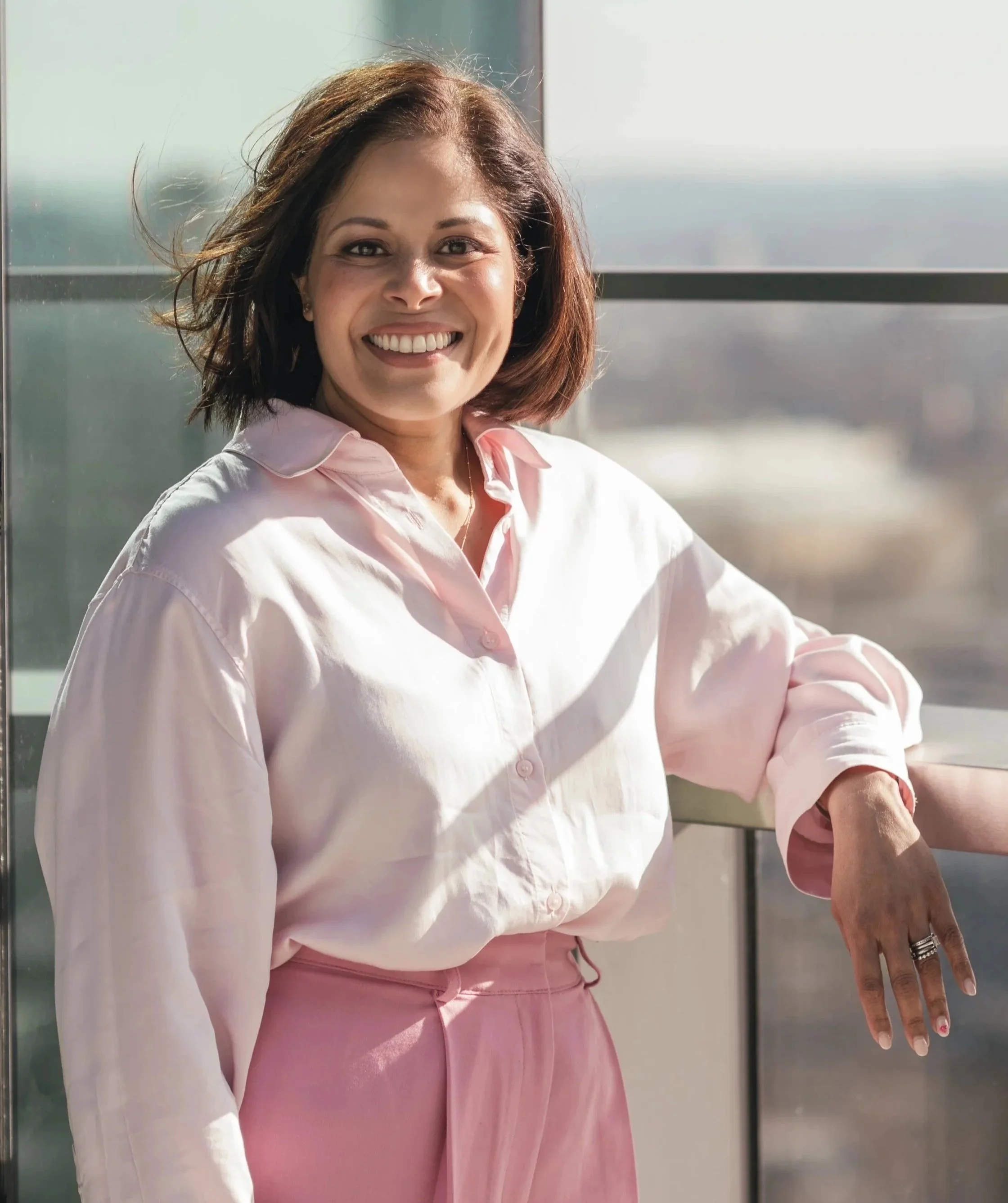 A woman with shoulder-length hair, smiling and wearing a light pink button-up shirt and pink pants, standing by a glass window with a cityscape in the background.