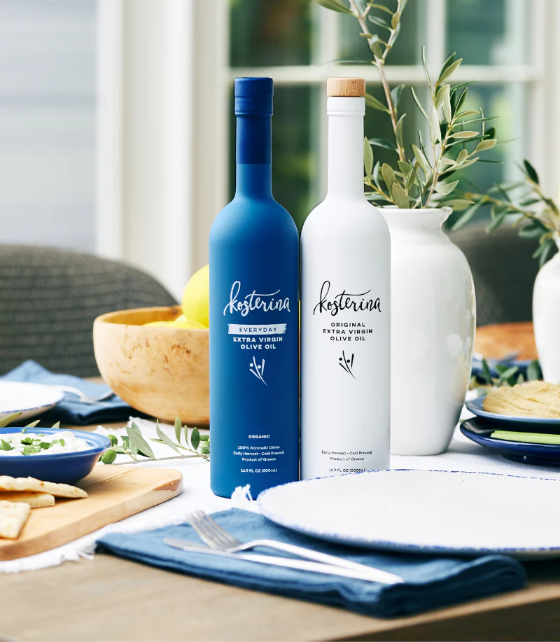 Two bottles of Kosterina extra virgin olive oil on a dining table, one blue and one white, surrounded by a bowl of lemons, a white vase with green leaves, plates, and silverware.
