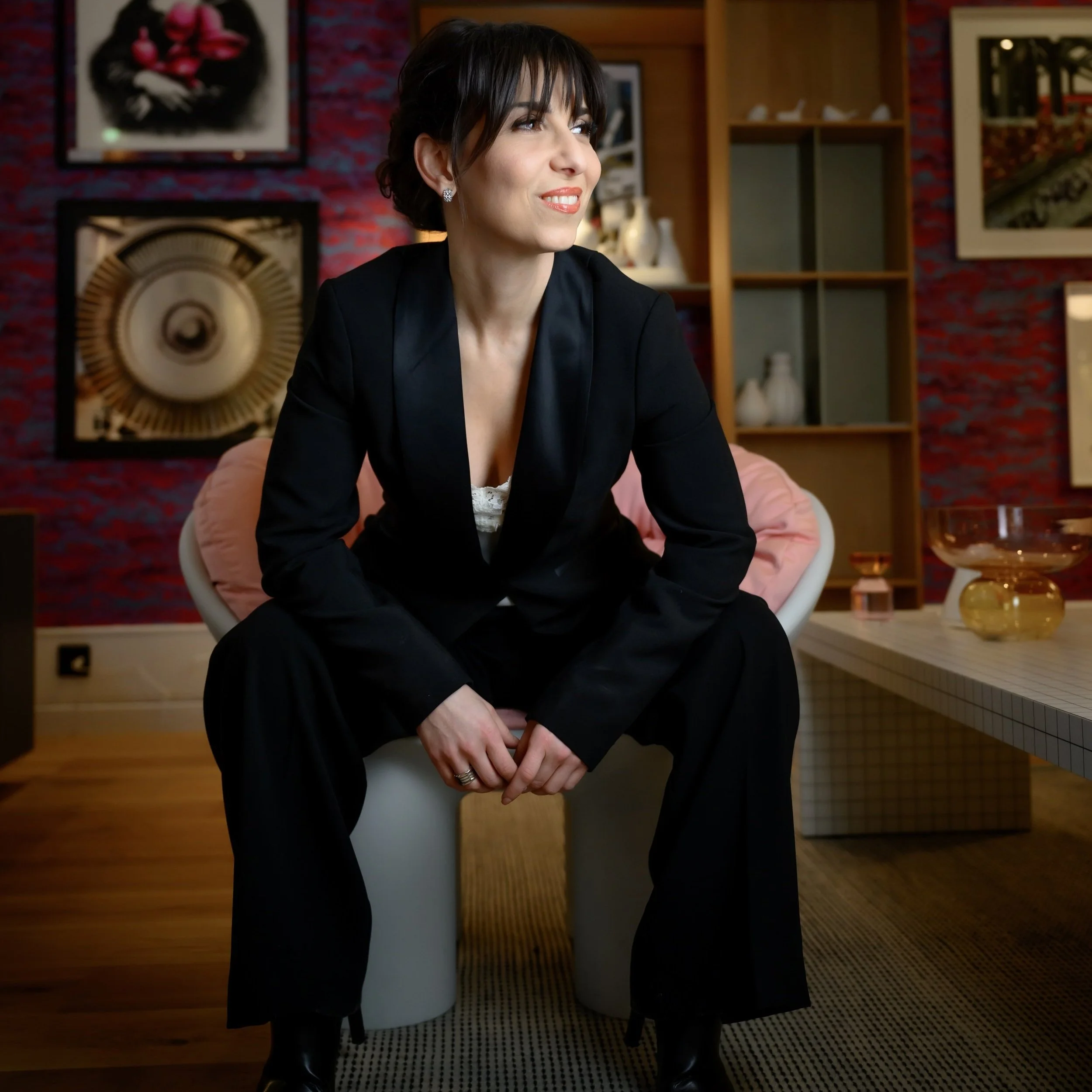 A woman sitting on a modern white chair in a stylish living room, wearing a black blazer and pants, with short dark hair and earrings, looking to the side with a smile.