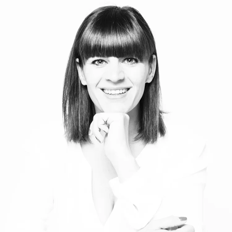 A smiling woman with shoulder-length dark hair and bangs, resting her chin on her hand, posed against a white background.