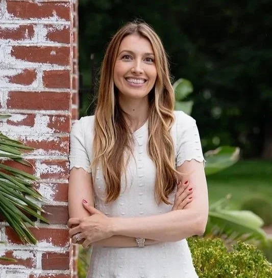 A smiling woman with long wavy hair standing outdoors near a brick wall with green plants and trees in the background.