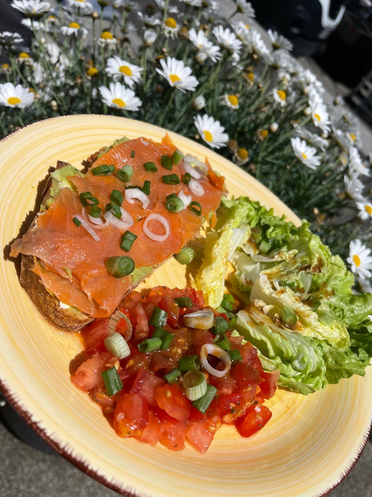Open-faced smoked salmon sandwich with green onions, along with fresh tomato and lettuce salad, on a yellow plate outside with daisies in the background.