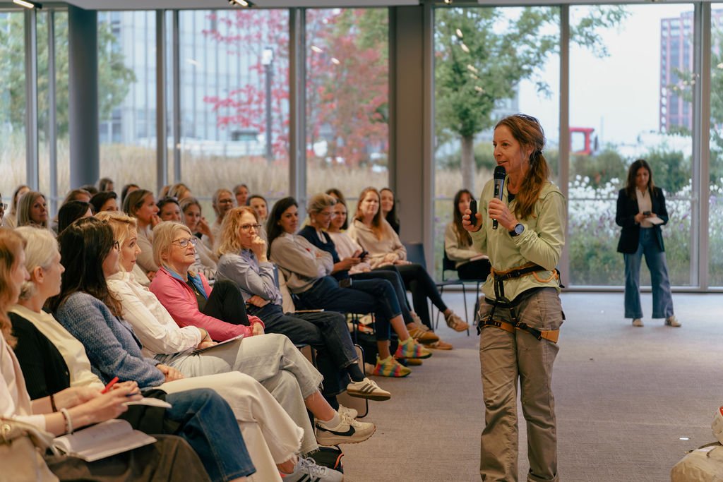 A woman giving a presentation or speech to an audience of women seated in a spacious room with large windows showing outdoor trees and buildings.