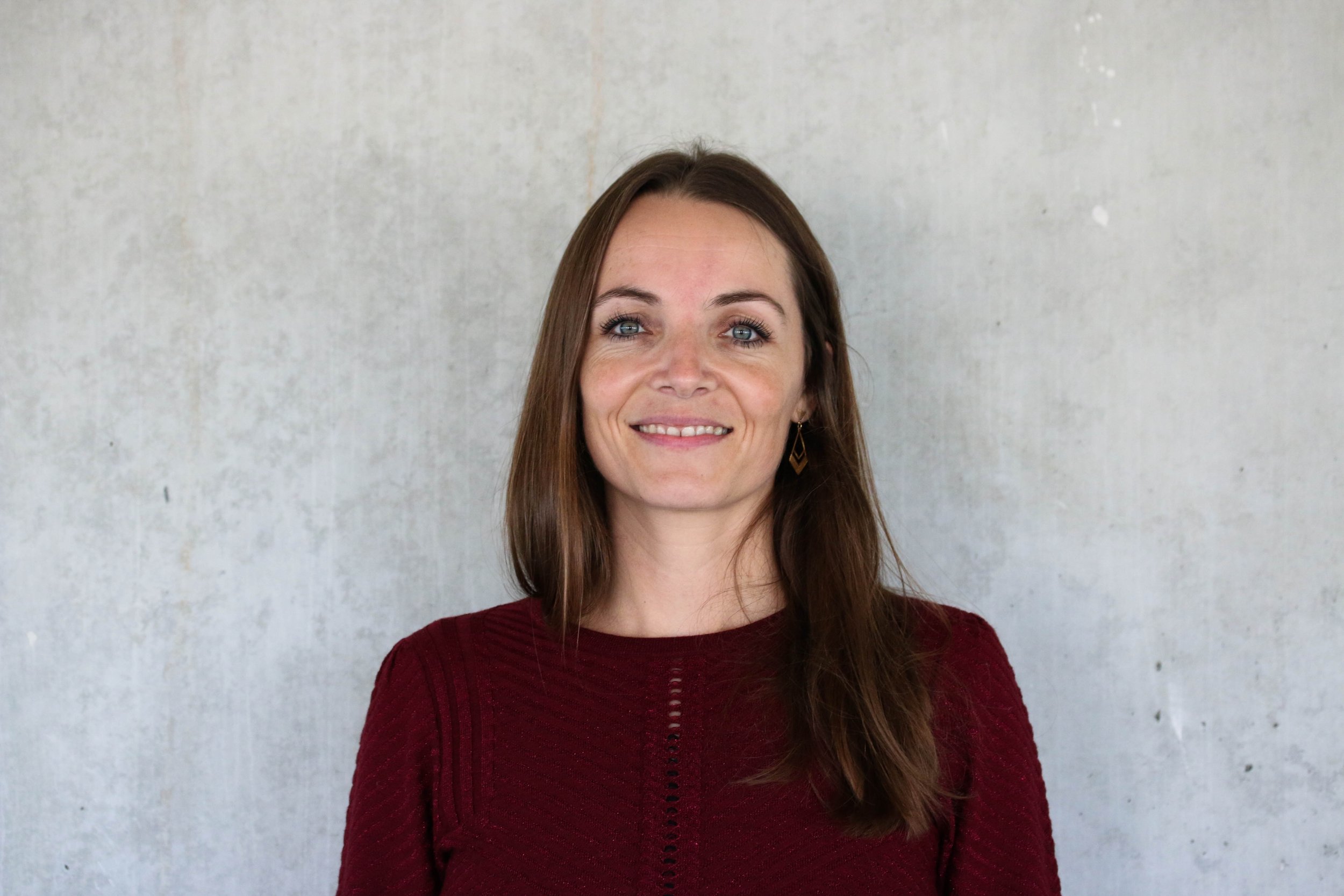 A woman with long brown hair and blue eyes smiling, wearing a red sweater, standing against a plain, gray concrete wall.