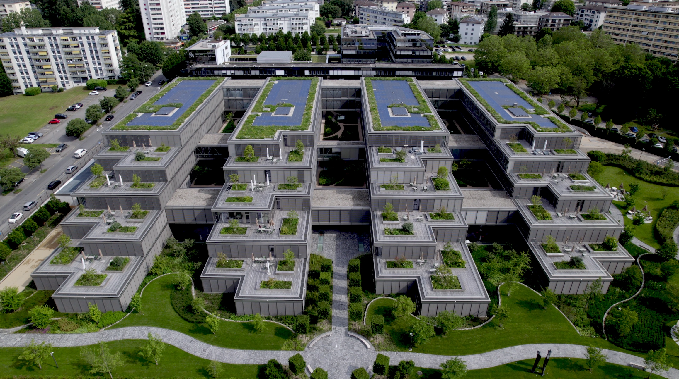 An aerial view of a modern, multi-story apartment complex with rooftop gardens and solar panels, surrounded by greenery and other residential buildings.