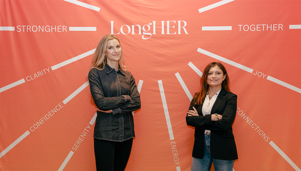 Two women standing in front of a red backdrop with words like 'LongHER', 'Stronger', 'Together', and 'Joy.' They are crossing their arms.