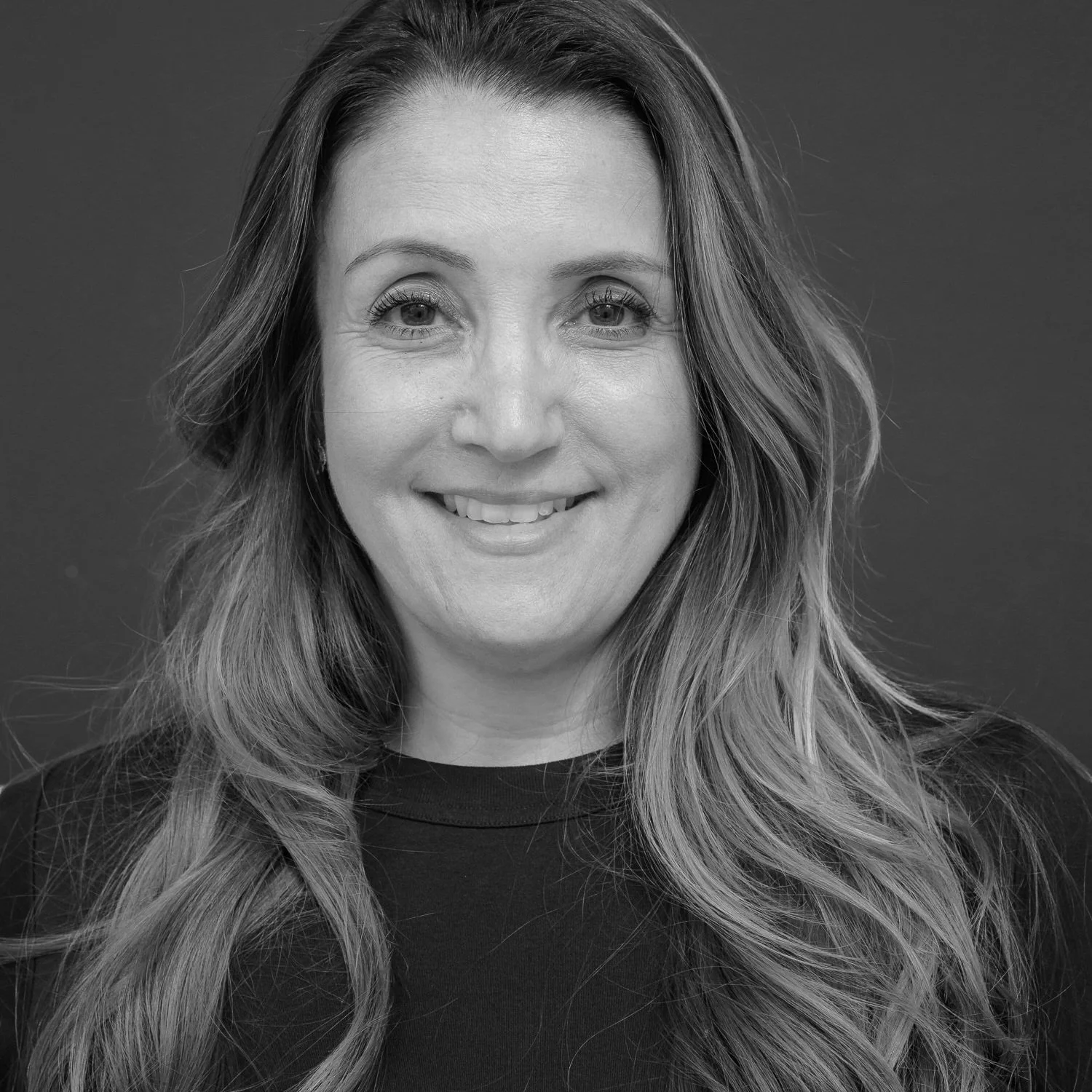 Black and white portrait of a smiling woman with long wavy hair, wearing a dark top, against a plain background.