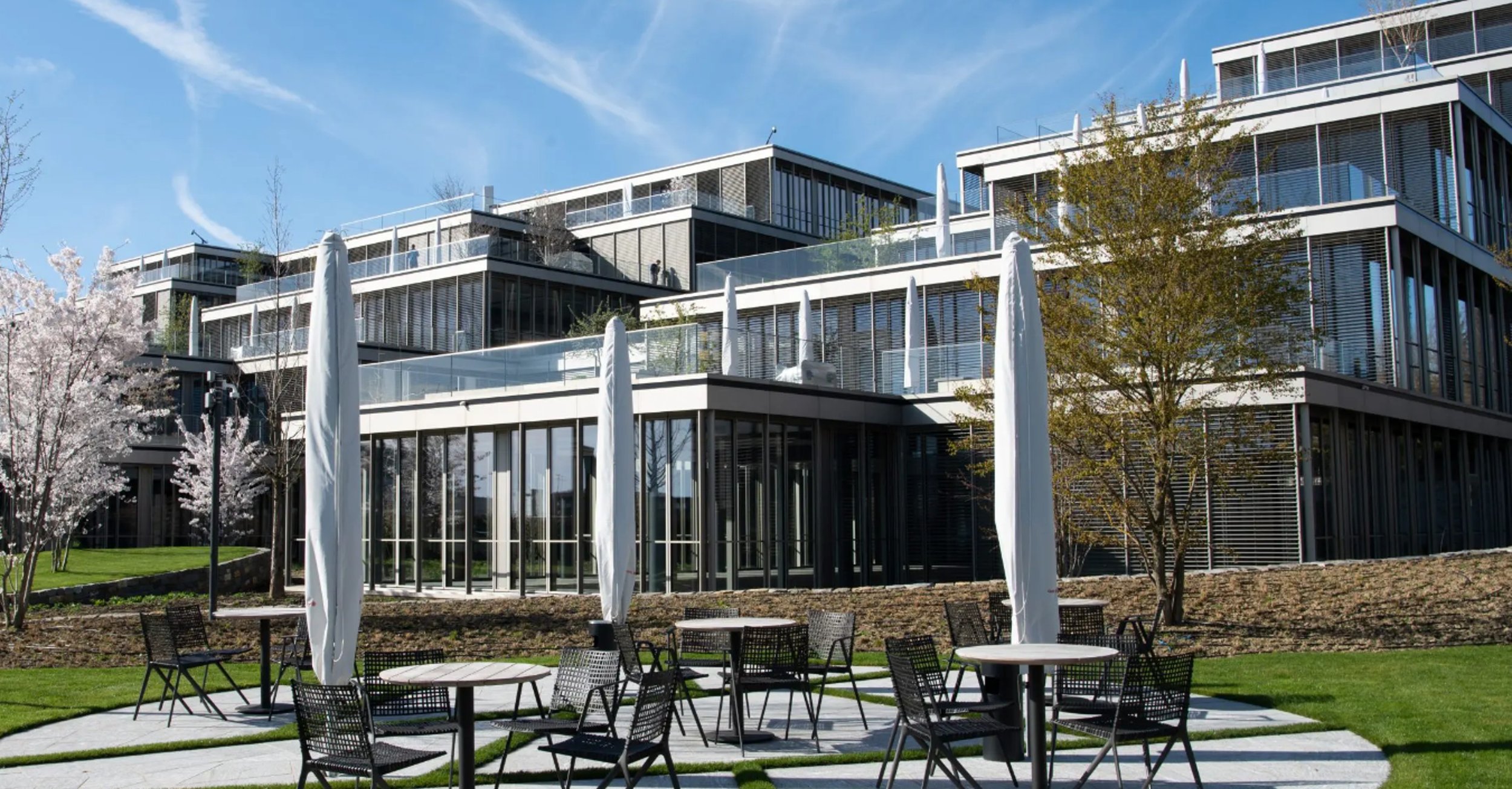 Modern multi-story building with glass exterior, outdoor seating area with tables and chairs, umbrellas, trees, and a well-maintained lawn under a blue sky.