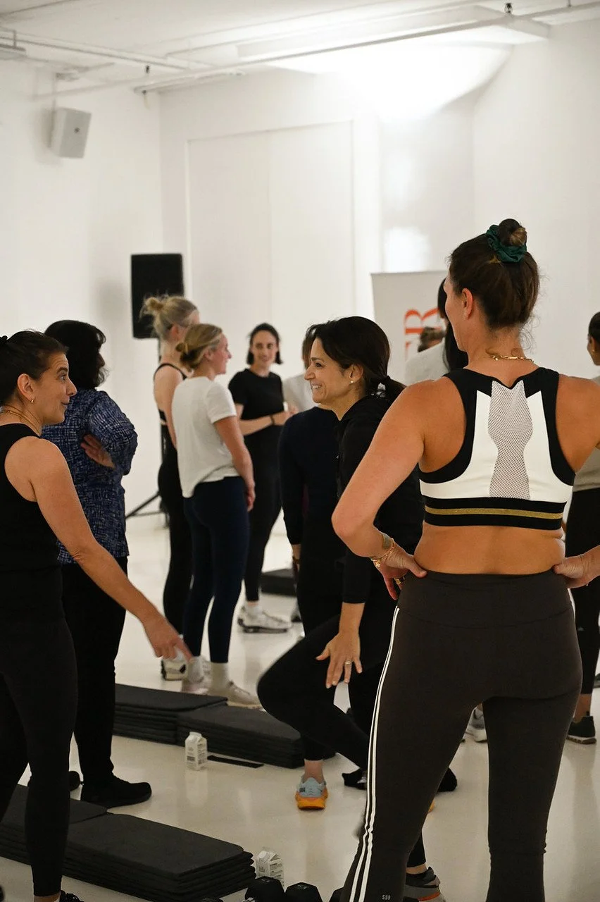 Group of women in athletic clothing gathered in a fitness studio, engaging in conversation and stretching.