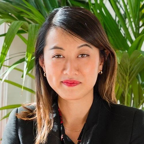 A woman with straight hair, wearing a black blazer and gold earrings, sitting in front of green plants.