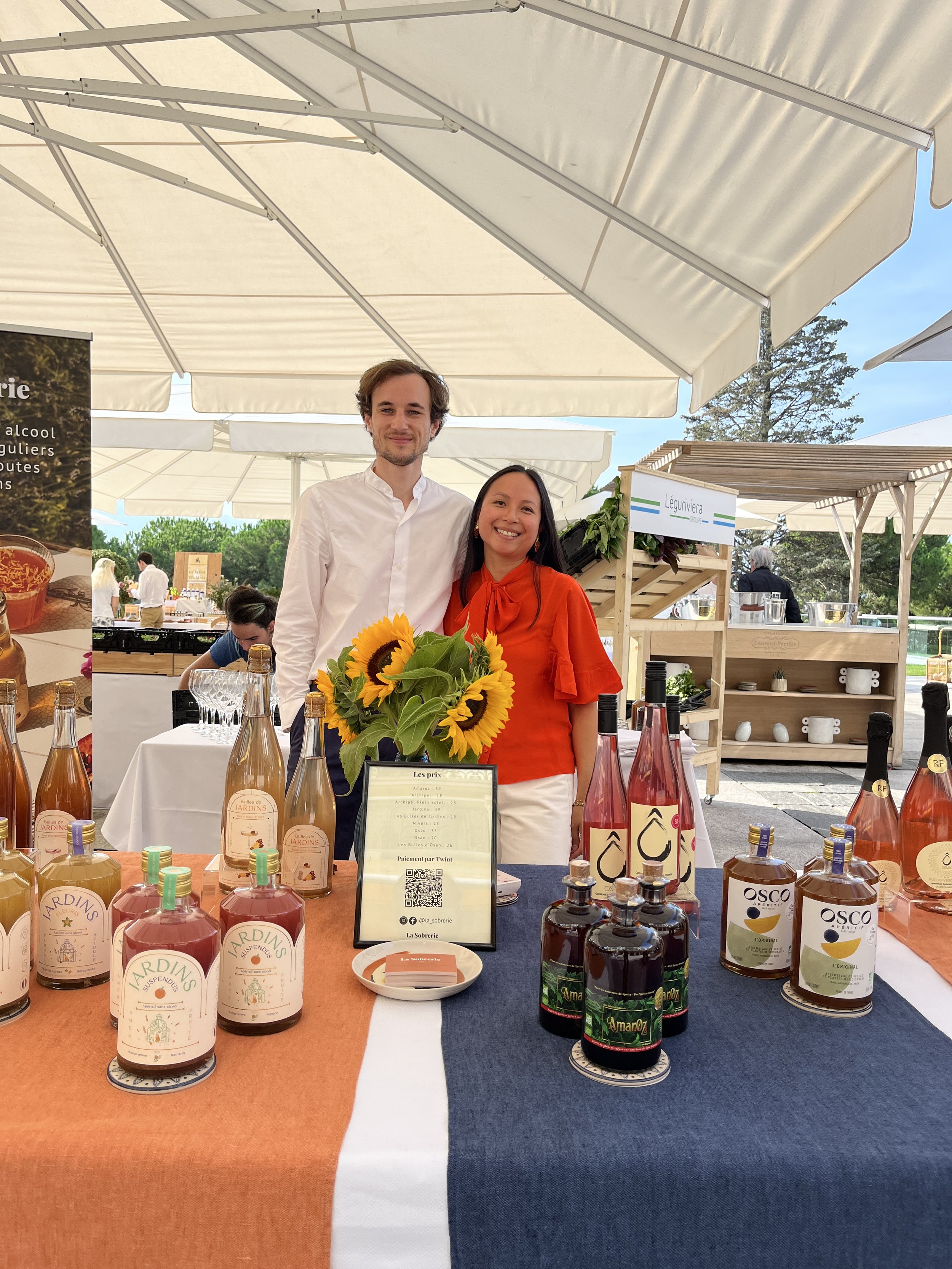 Two people standing behind a table with bottles of beverages at an outdoor market or fair, with white tents in the background and a clear sky.
