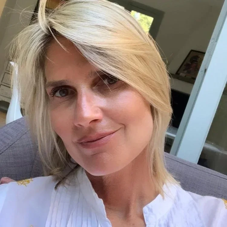 A woman with blonde, shoulder-length hair and fair skin is smiling and looking at the camera inside a room with a window behind her.