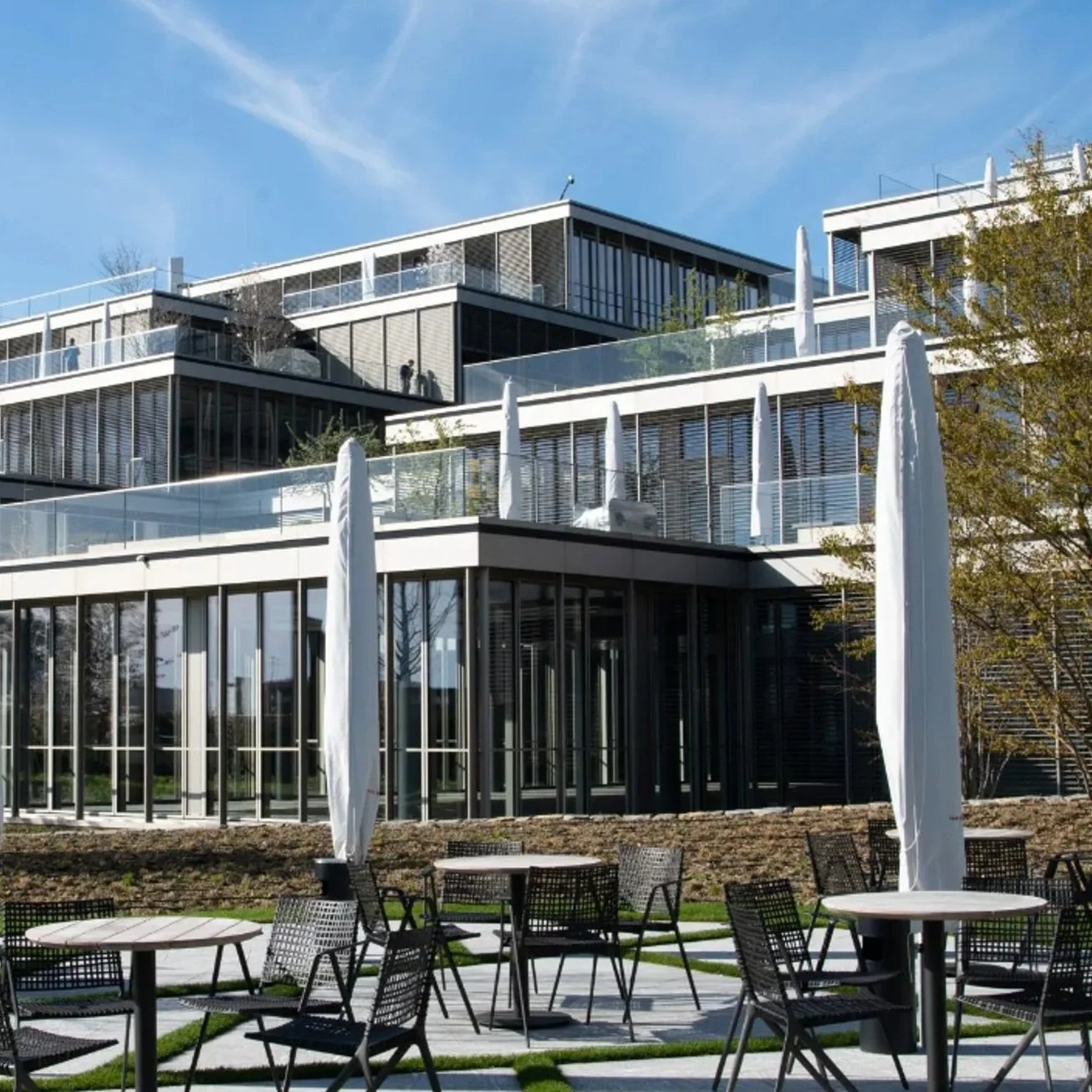 Modern multi-story building with glass windows and balconies, outdoor patio area with tables and chairs, and parasols, under a clear blue sky.