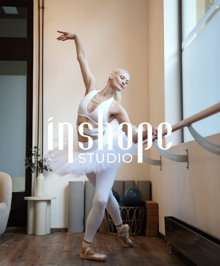 A ballerina in a white tutu and leotard practicing at a ballet barre in a dance studio, with large windows and exercise equipment in the background.