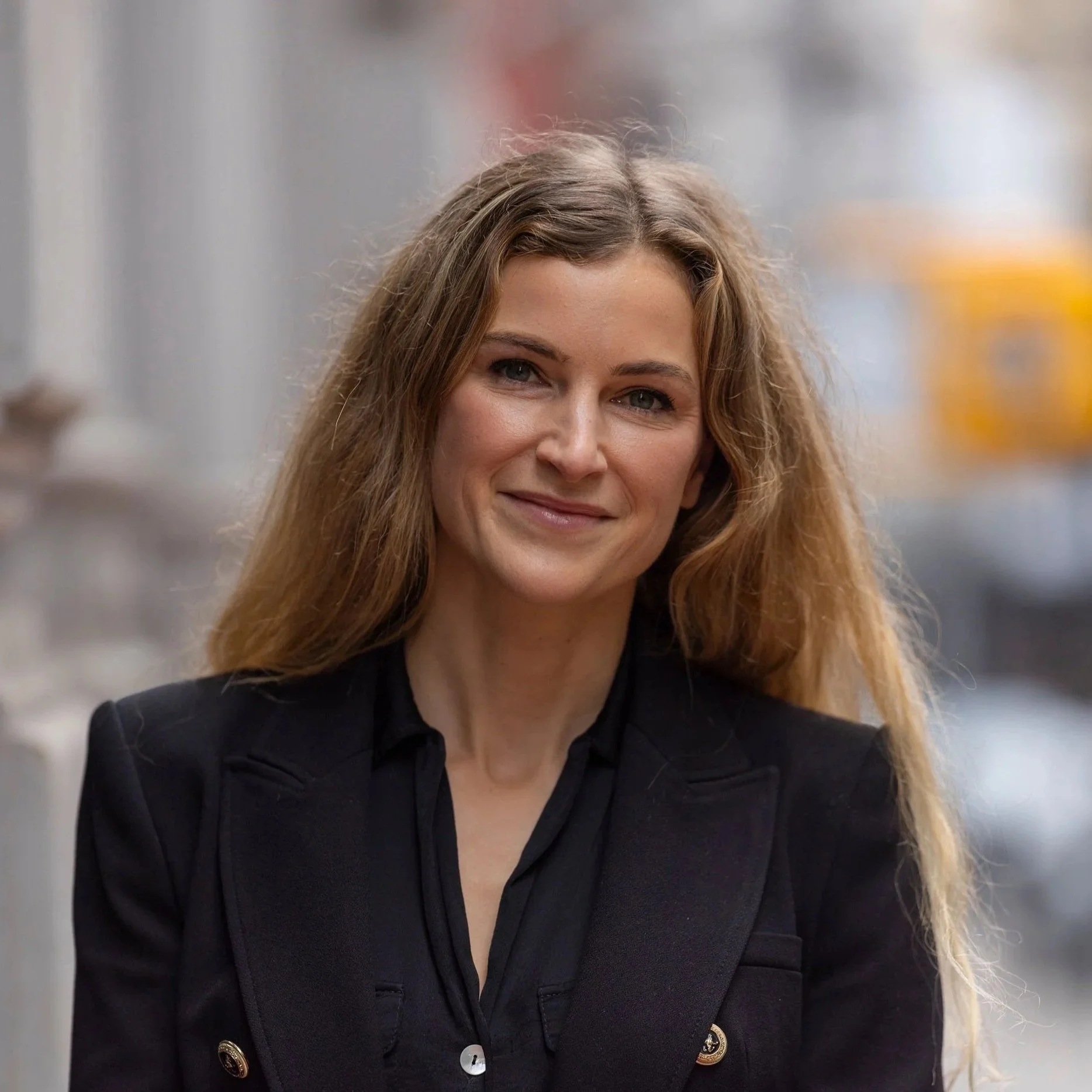 A woman with long, wavy blonde hair, wearing a black blazer over a black top, standing outdoors in an urban setting.