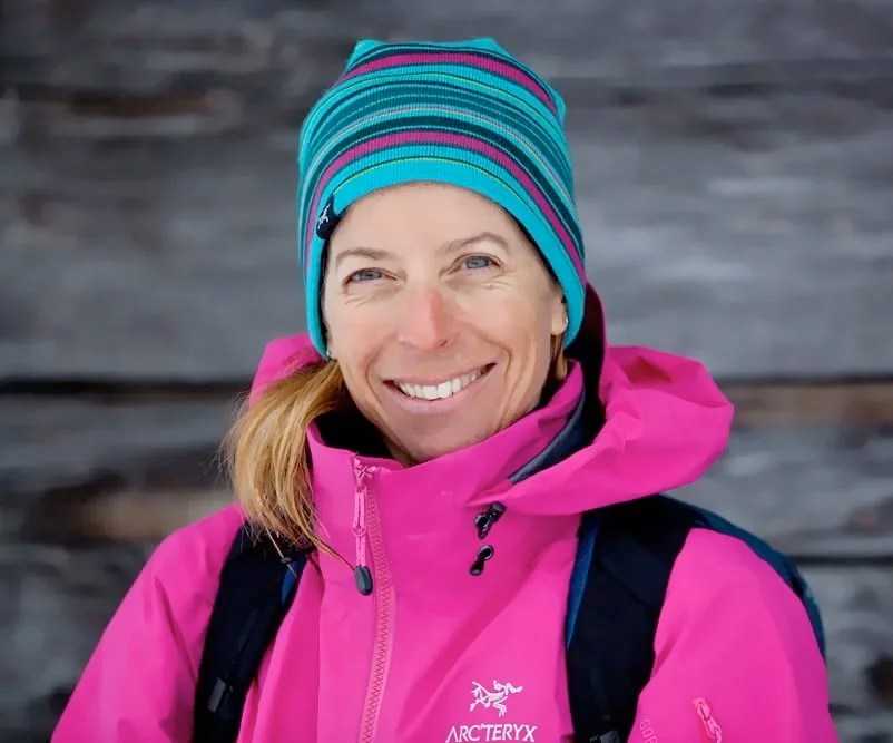 Woman in pink outdoor jacket and colorful striped beanie smiling at camera with a wooden background.