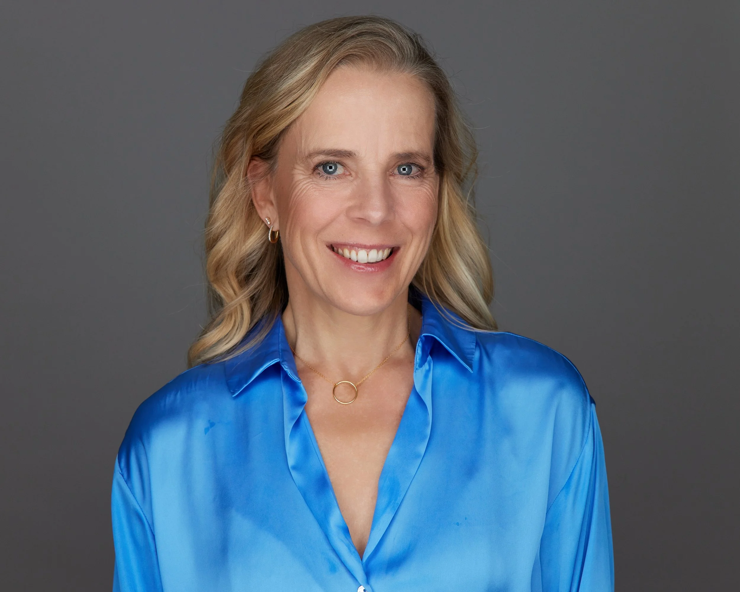 A smiling woman with blonde hair, wearing a shiny blue silk blouse and small hoop earrings, posed against a gray background.