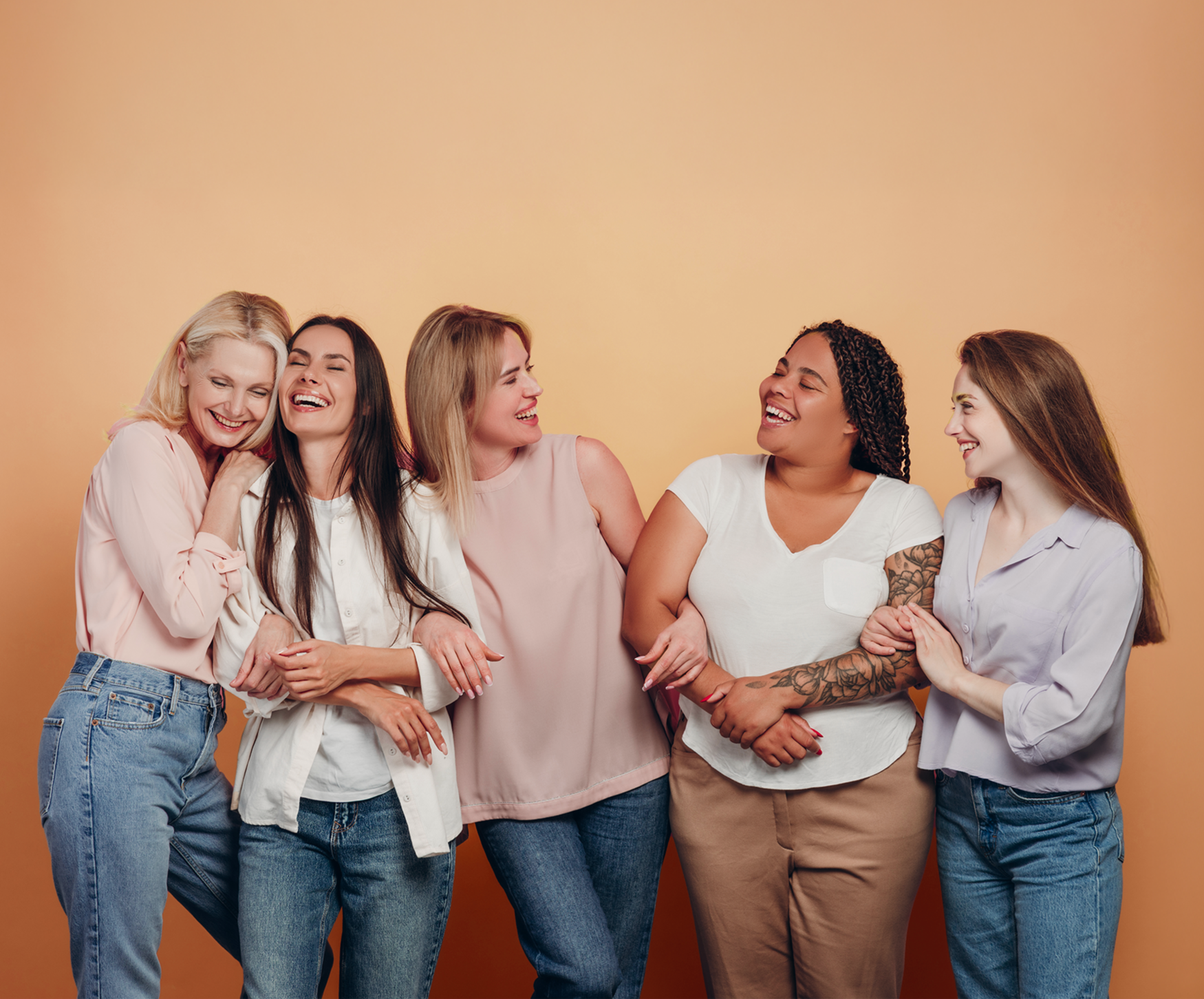 Group of five women standing close together, smiling, and interacting affectionately against a warm orange background.