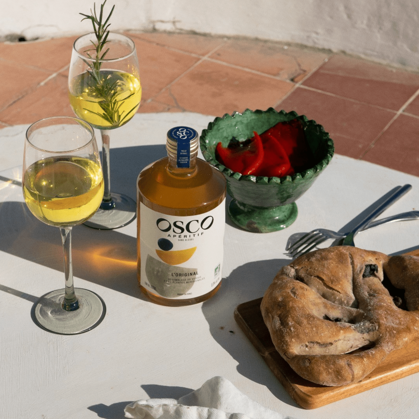 A table outdoor with two glasses of white wine, a bottle of Osco Aperitif, a bowl of red peppers, a baked pastry with chocolate chunks on a wooden board, and a fork and knife.