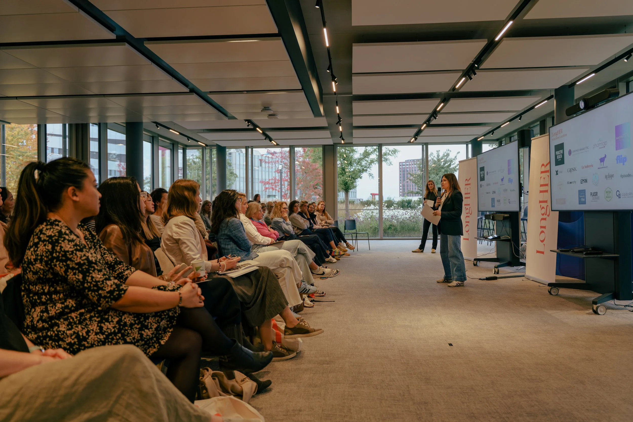 Women presenting to an audience in a modern conference room with large windows, multiple screens, and banners reading "LONGHIER." The audience is attentively listening, seated in rows, with some taking notes.