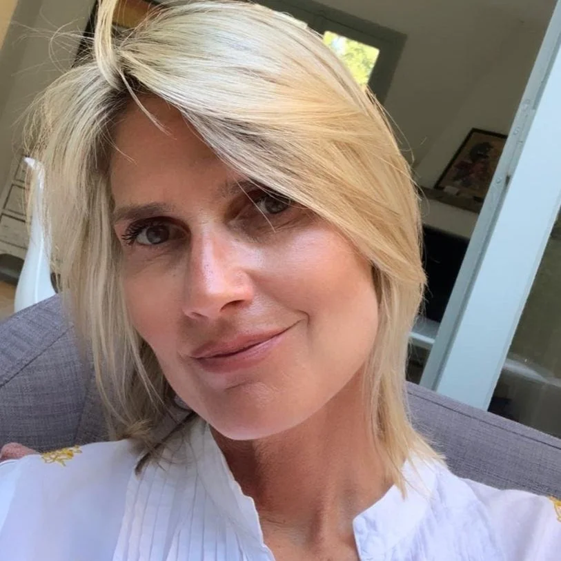 A smiling woman with blonde hair, wearing a black top with a blue detail, sitting in a room with a wooden dresser and a bedding visible in the background.