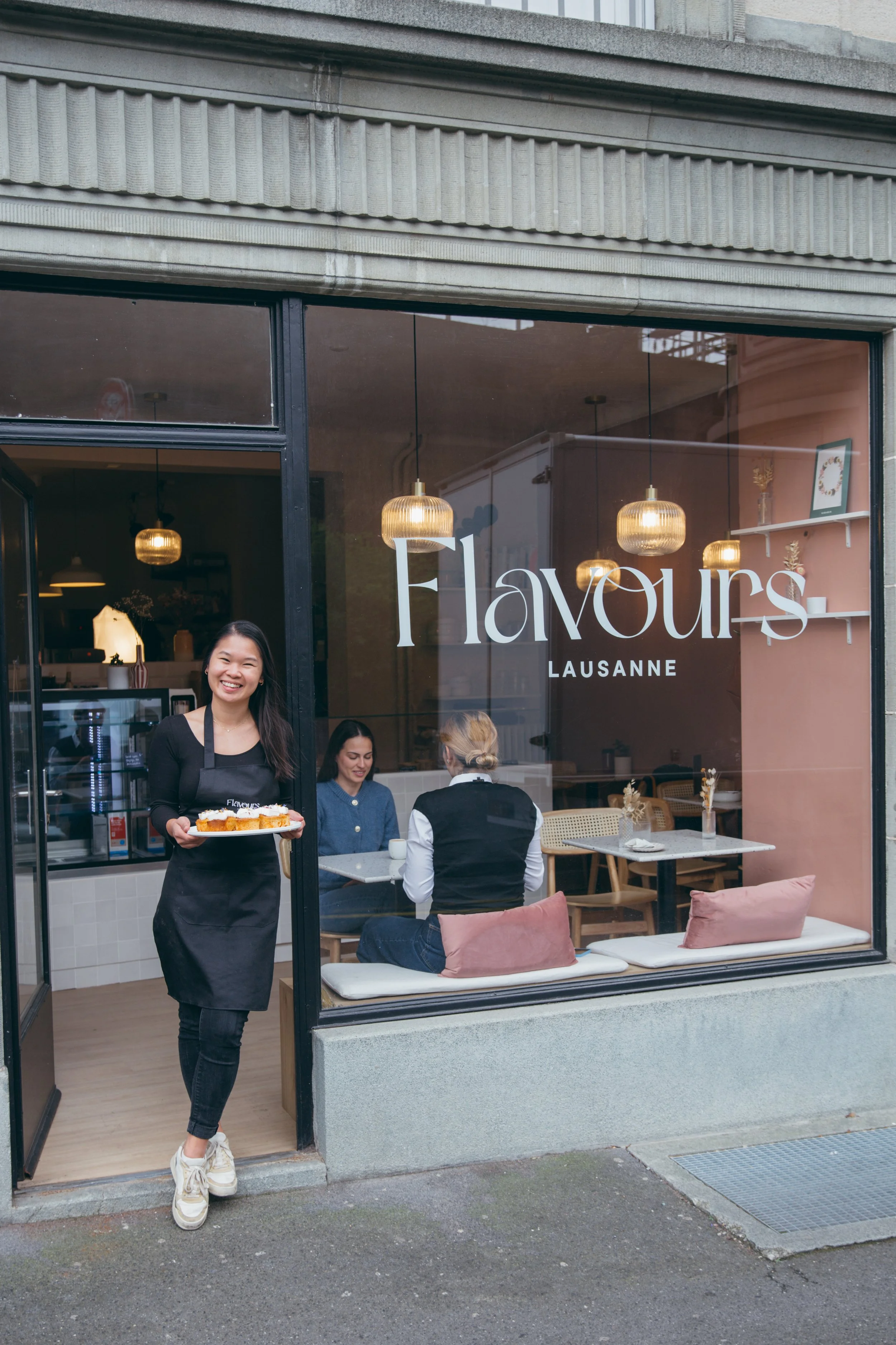 Smiling waitress holding a tray with food outside a cafe named Flavours Lausanne with customers seated inside.