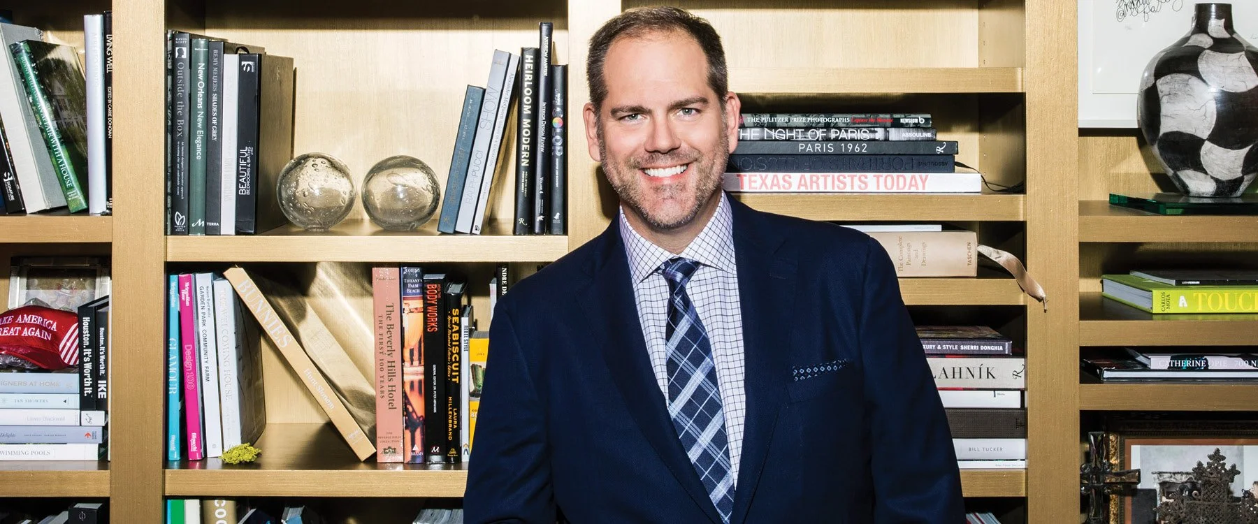 A man in a navy suit and patterned shirt, smiling, standing in front of a bookshelf with various books and decorative items.