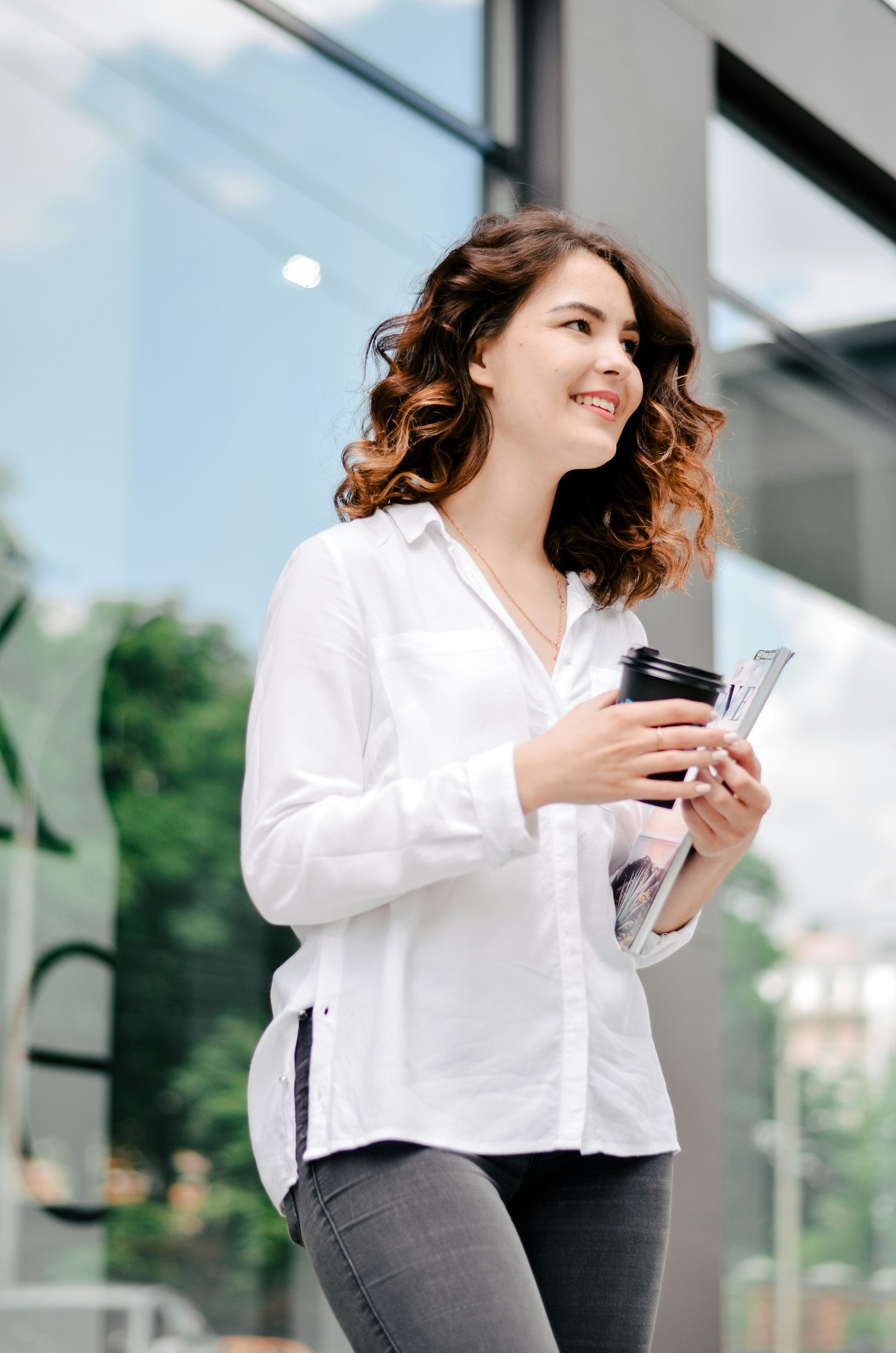 Young woman with curly hair smiling, holding a travel coffee cup and a magazine, standing outside a modern building considering egg freezing