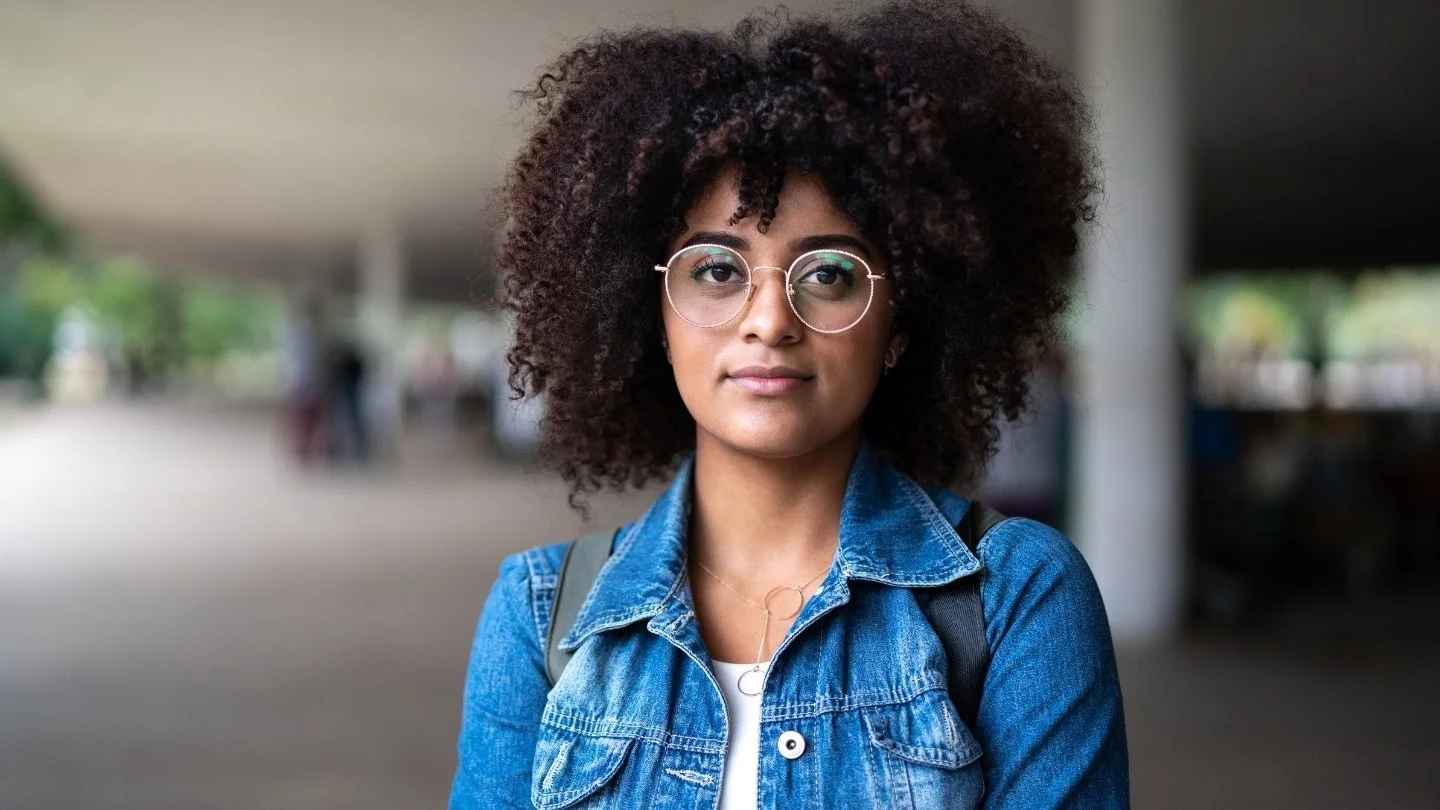 Young woman with curly hair, wearing glasses and a denim jacket, standing outdoors with blurred background.