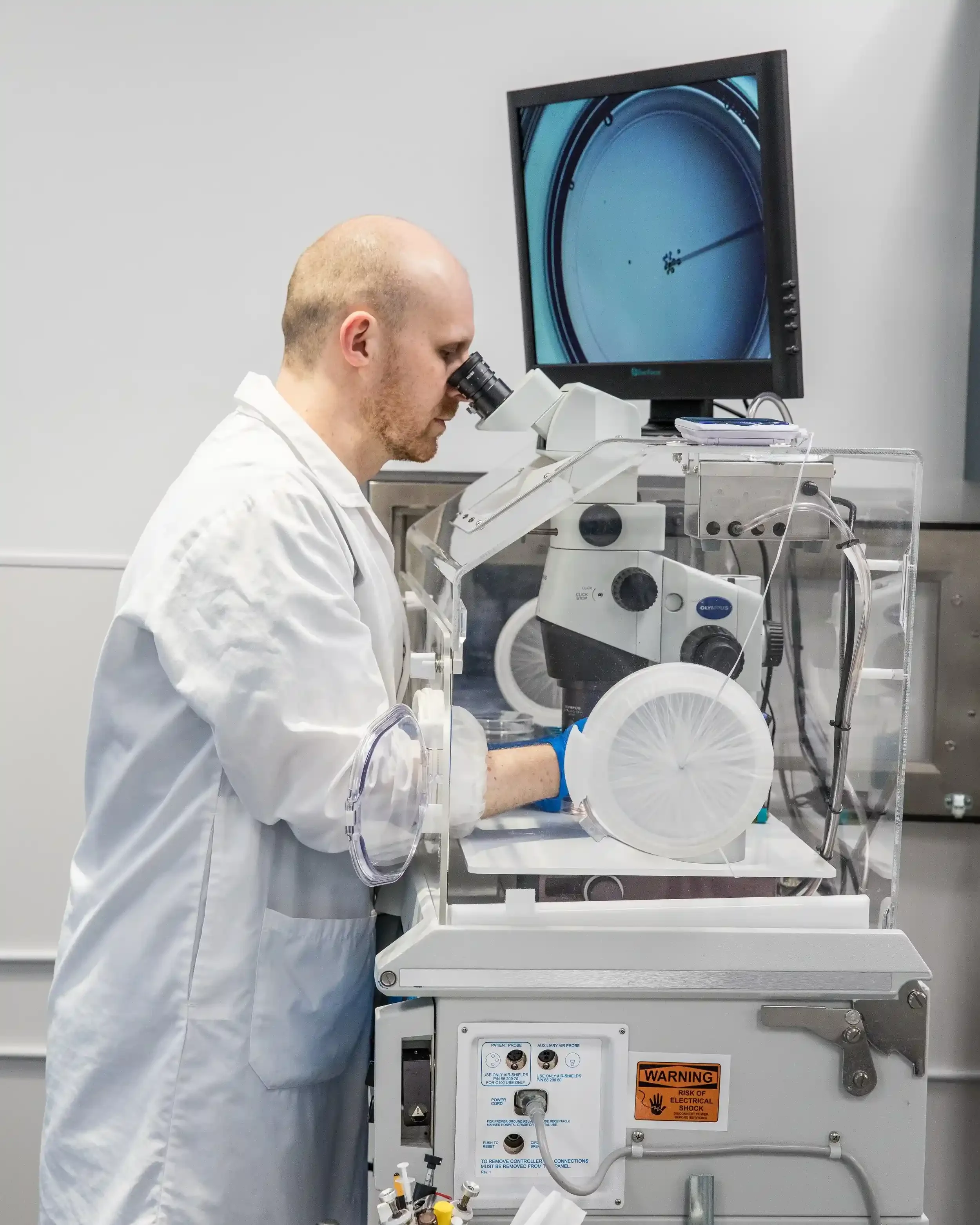 A scientist wearing a white lab coat and blue gloves looking through a microscope in an IVF laboratory at CCRM Toronto.