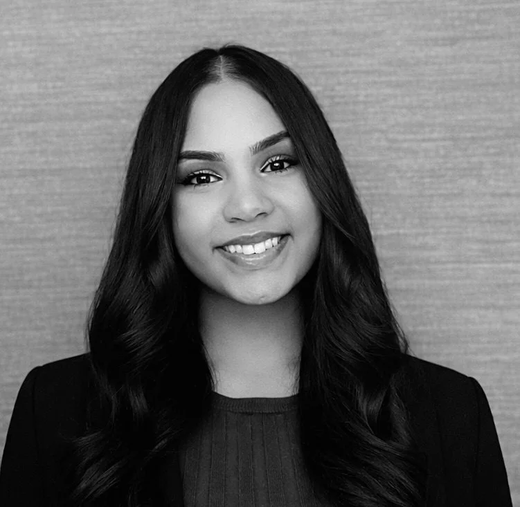 Black and white portrait of a young woman with long wavy hair smiling, wearing a dark jacket and blouse, standing in front of a textured wall.