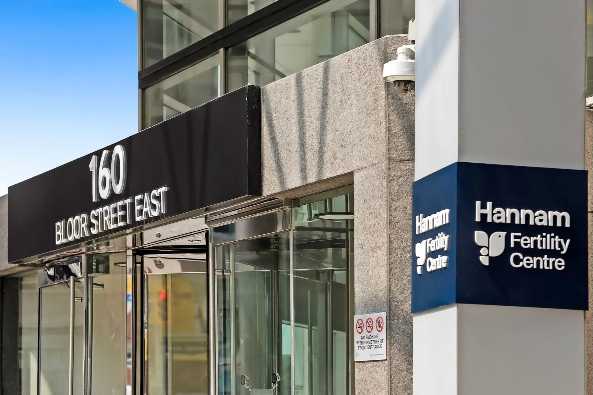 Entrance of  160 Bloor Street East building. The building has glass doors and windows, with a security camera mounted on the exterior wall. A blue sign indicates the location as the 'Hannam Fertility Centre.'