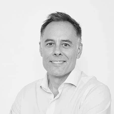 A black-and-white portrait of a man with short hair, smiling, wearing a white shirt, against a plain light background.