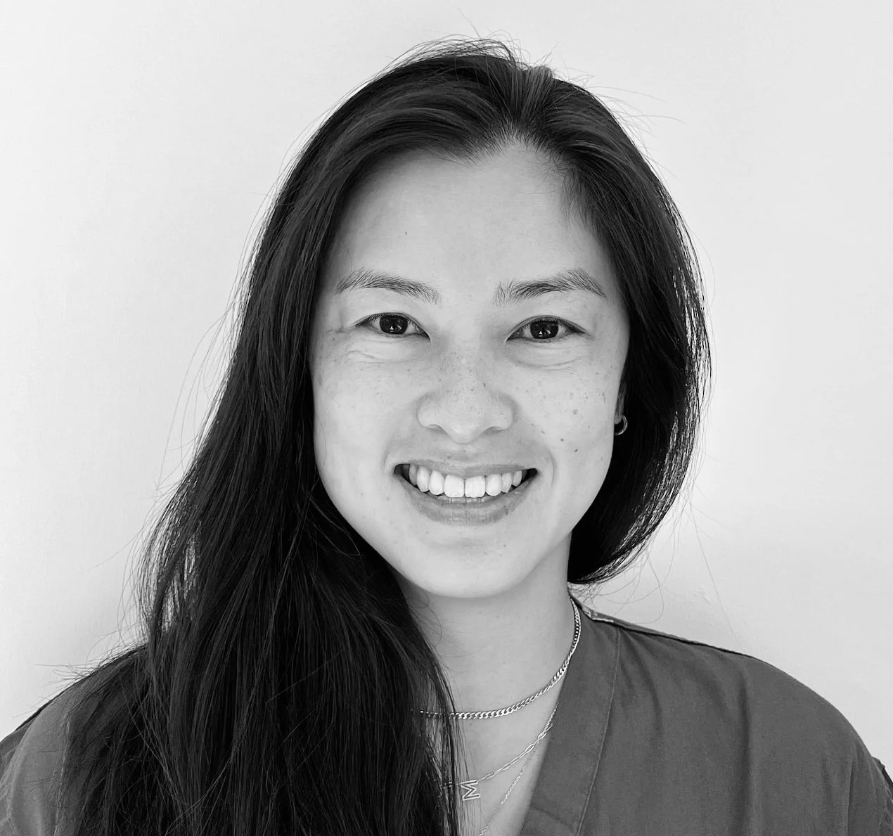 Black and white portrait of a smiling woman with long dark hair, wearing a necklace and a dark top, against a plain light background.