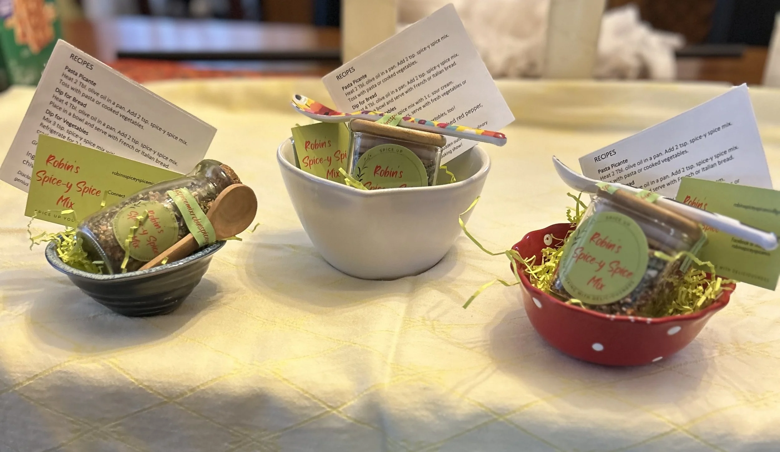 Gift basket items: Round and heart-shaped bowls with various products: spice mix in round or square jars, with recipe cards and serving spoons.