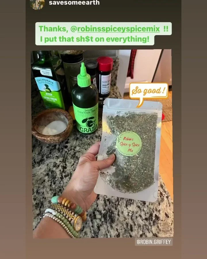 Hand holding a package of Robin’s Spice & Spice Mix in a kitchen with various bottles and containers on a granite countertop.