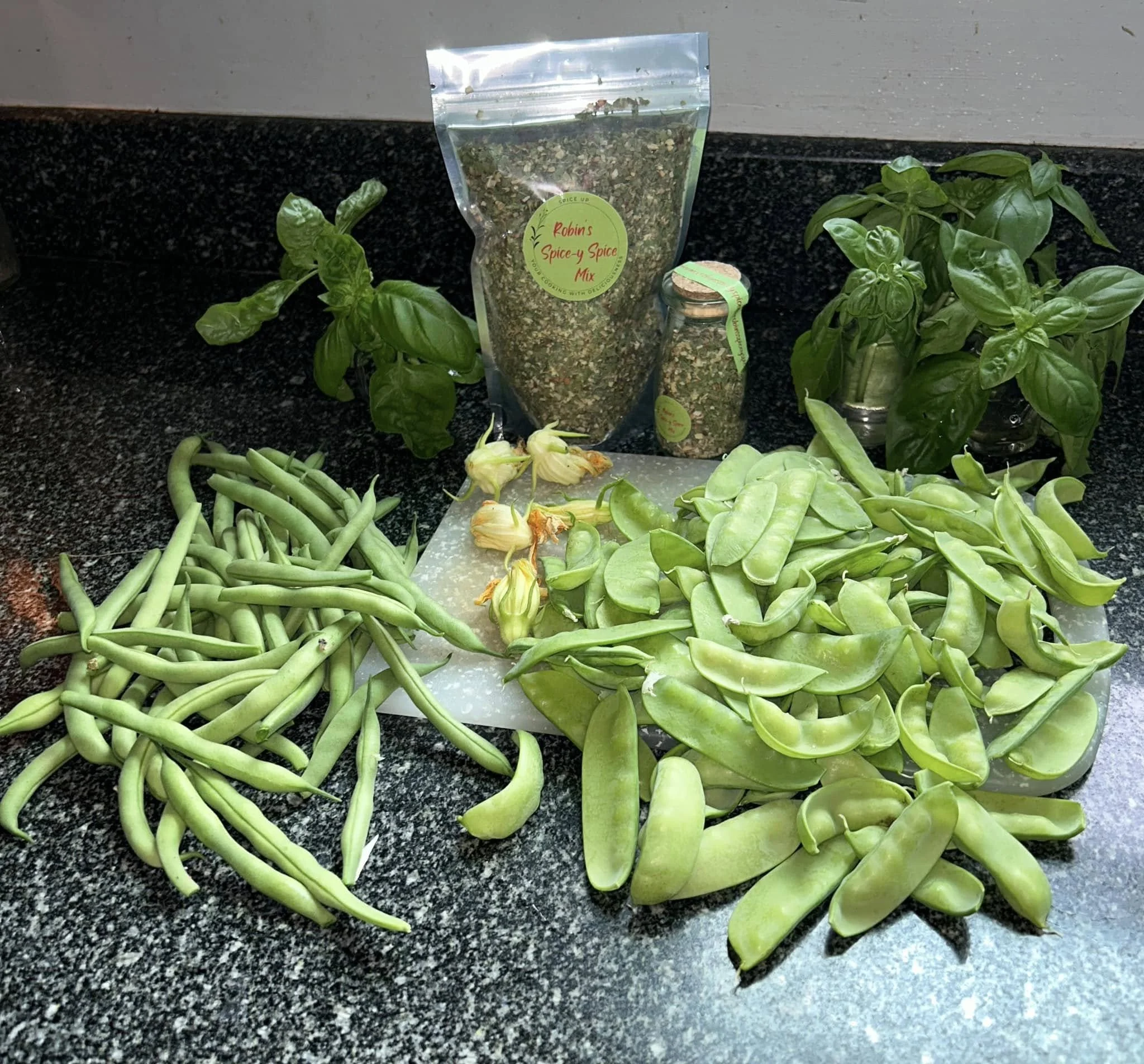 Fresh green beans, basil leaves, peas and a jar of dried herbs on a kitchen countertop, with a package labeled 'Robin's Spice y Spice Mix' in the background.