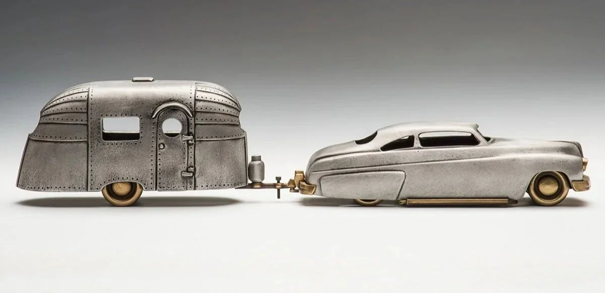 A vintage model of a teardrop trailer connected to a futuristic, streamlined car, both made of metal with gold accents.