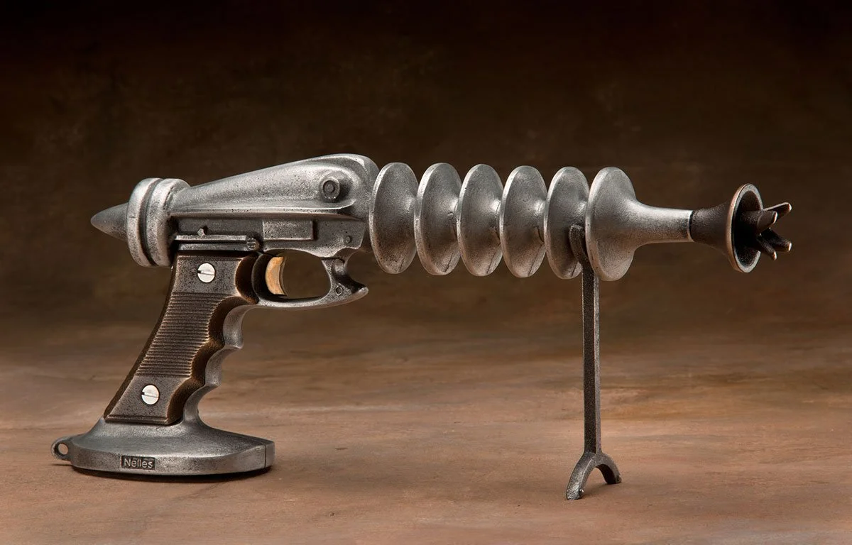 A sculpture resembling a handgun with a spiral, rocket-like tail, and a stand in place of the barrel, made of metallic material.