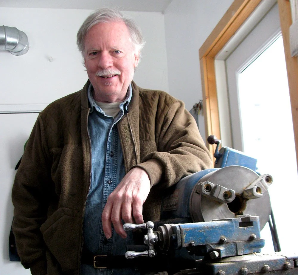 Scott Nelles, the owner of Nelles Studios, wearing a brown jacket over a blue shirt, standing next to a metal lathe in a workshop, smiling at the camera.