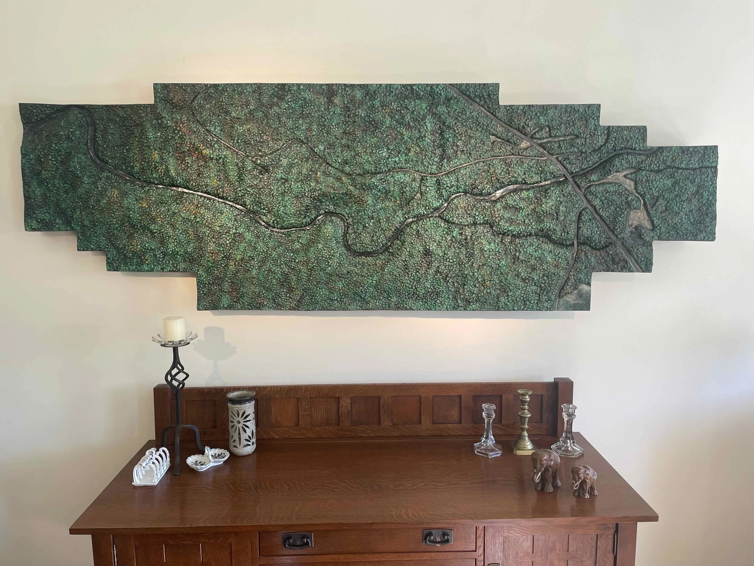 Decorative wall art resembling a map or landscape with green and brown hues, placed above a wooden sideboard with various decorative items including glass candlesticks, a ceramic elephant, and a candle holder.