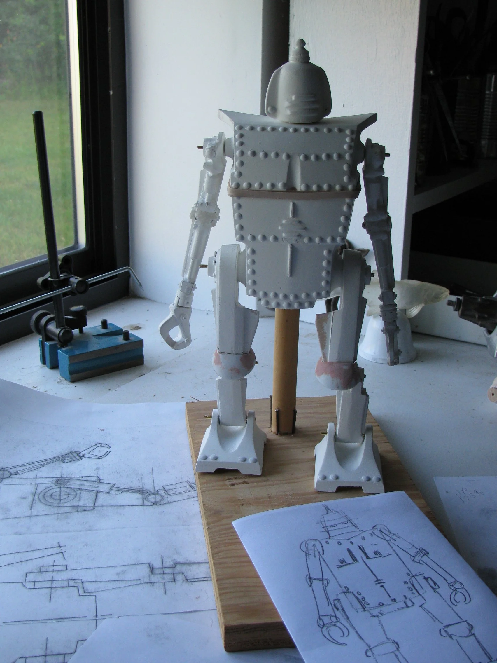 A white robot model with rivets, standing on a wooden base, positioned on a table near a window. There are technical drawings and sketches of robots and mechanical parts on the table.